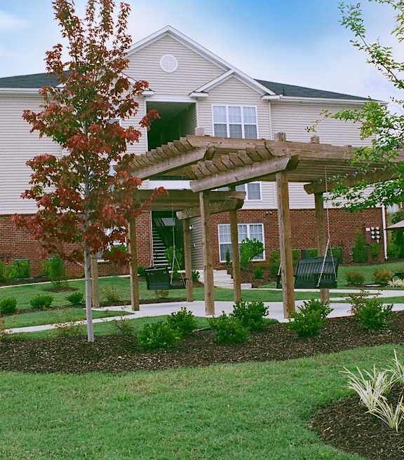 A building with a white exterior and a green awning is surrounded by a garden with a wooden pergola.