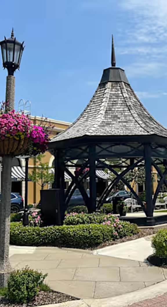 A gazebo with a black roof is surrounded by bushes and flowers.