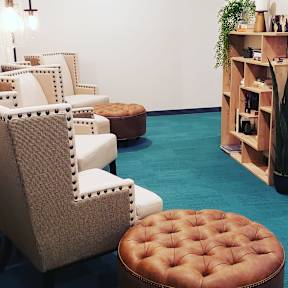 A brown tufted ottoman sits in front of a white chair with a wooden bookshelf in the background.