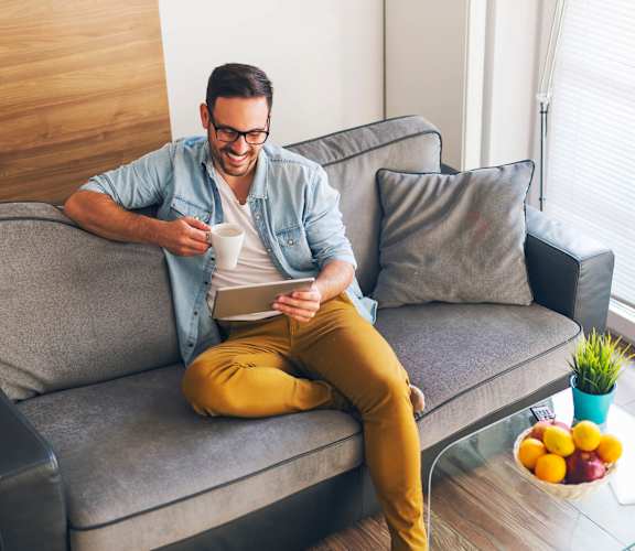 A man is sitting on a grey couch with a cup of coffee in his hand at 26FIFTY Apartments, Berkeley 94704