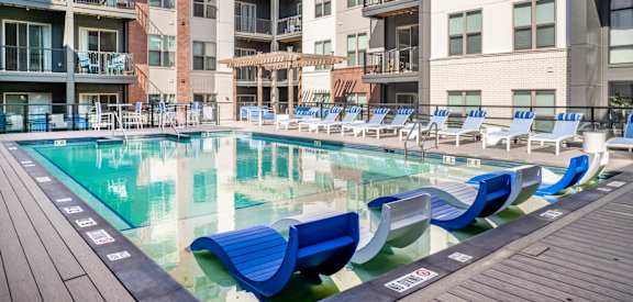 A pool with blue lounge chairs and a wooden deck in front of apartment buildings.