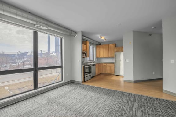 an empty living room with a large window and a kitchen in the background