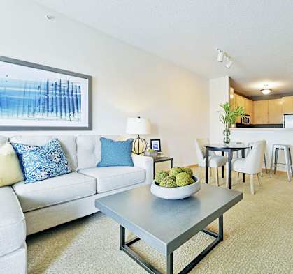 A modern living room with a white couch, a grey coffee table, and a dining area in the background.