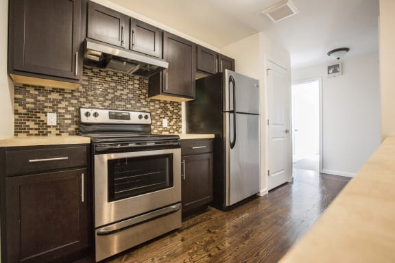 Kitchen with stainless steel appliances, hardwood floors, dark wooden cabinets, off white counter tops, and black and white tile patterned back splash