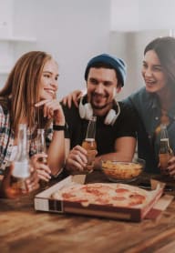 group of young people sitting around a table eating pizza and drinking beer