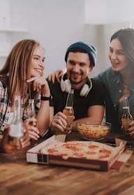 group of young people sitting around a table eating pizza and drinking beer