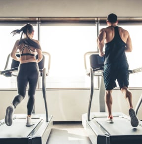 Man and woman running on treadmills in a gym with large windows.