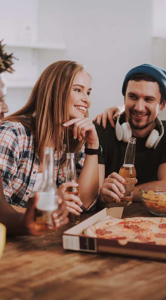 A group of people are gathered around a table with a bowl of flour, a pizza, and glasses of beer.