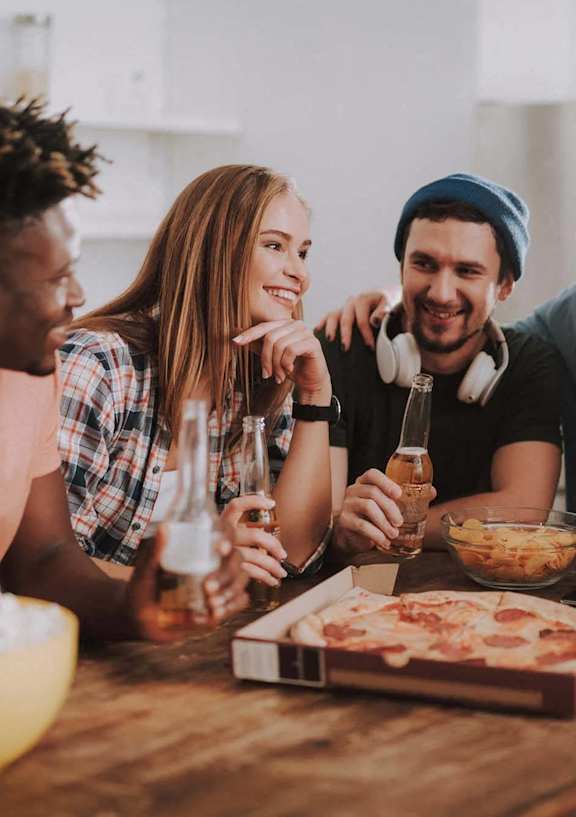 group of young people sitting around a table eating pizza and drinking beer