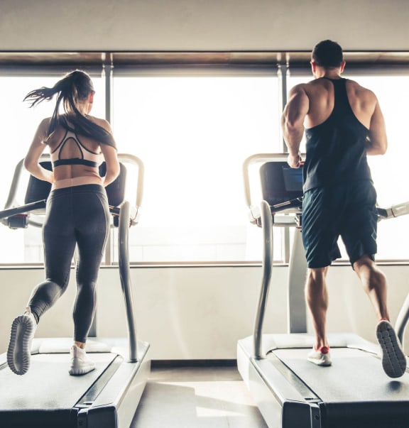 Man and woman running on treadmills in a gym with large windows.