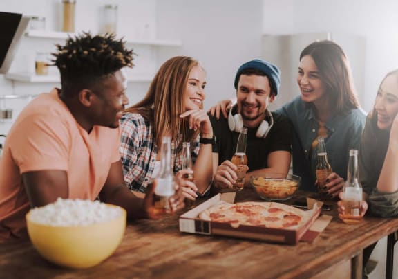 group of young people sitting around a table eating pizza and drinking beer