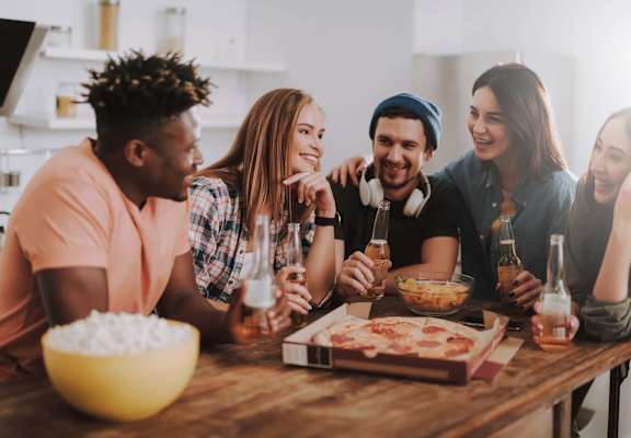 A group of people are gathered around a table with a bowl of flour, a pizza, and glasses of beer.
