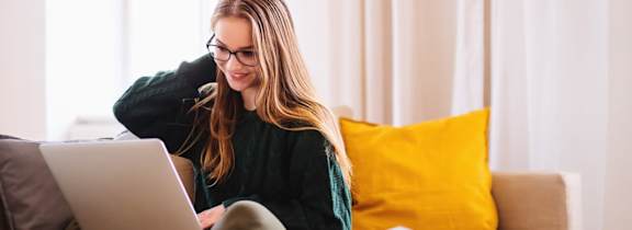 A woman with glasses is sitting on a couch using a laptop.