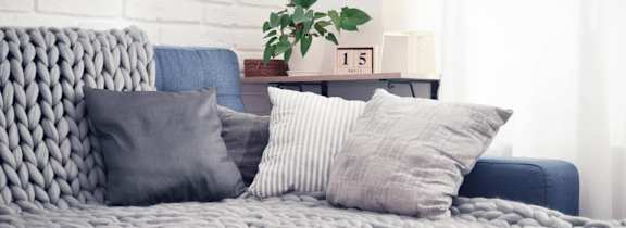 A grey knitted couch with a blue chair and a small table with a plant and a box on it.