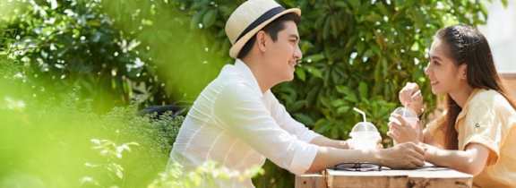 A man in a hat is sitting at a table with a woman, both are holding drinks.