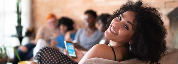 A woman with curly hair is smiling and holding a phone in a room with other people in the background.