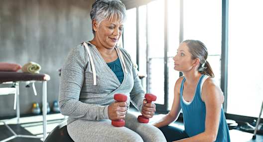 a woman sitting on an exercise ball with a woman standing next to her holding dumbbells