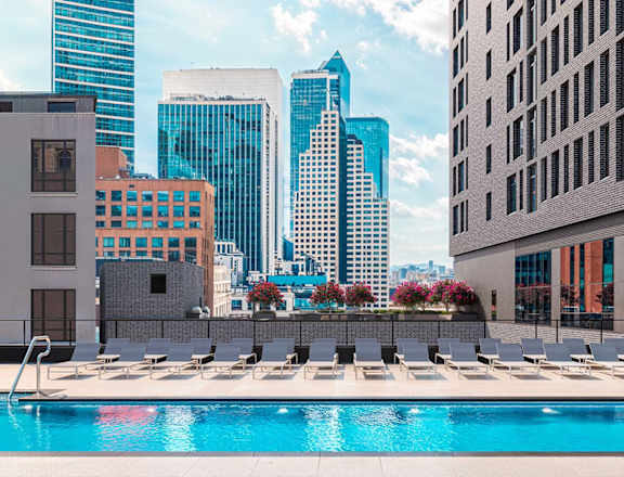 A swimming pool with sun loungers in the foreground and a city skyline in the background.