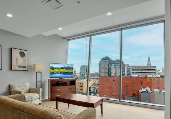 A living room with a tan couch, a coffee table, and a large window with a view of the city.