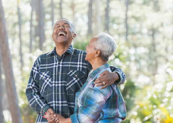 An older couple is standing in a forest, holding hands and laughing together.