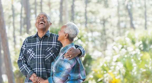 An older couple is standing in a forest, holding hands and laughing together.