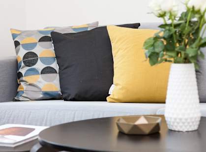 A grey couch with a yellow pillow and a vase with white flowers on a table.