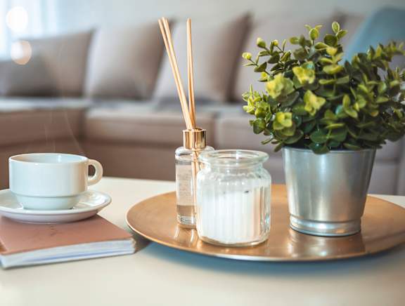 A white cup and saucer are on a table with a notebook, a candle, and a plant.