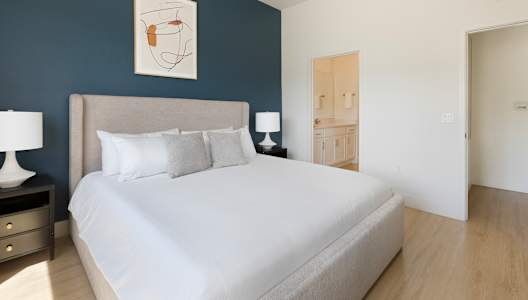 A bed with a white sheet and two pillows sits in a room with a blue wall.