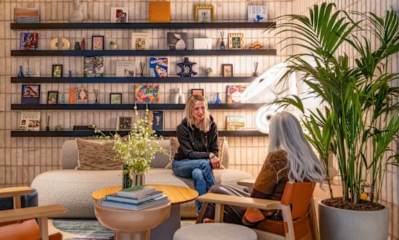 A woman sits on a chair in a room with a shelf full of books and decorative items.