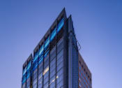 Thumbnail 1 of 18 - A tall building with blue glass windows stands in the evening sunlight.