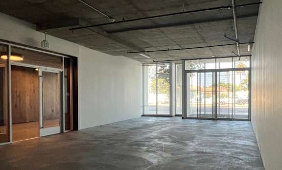 A large, empty room with a concrete floor and ceiling, and a view of the outside through large windows.