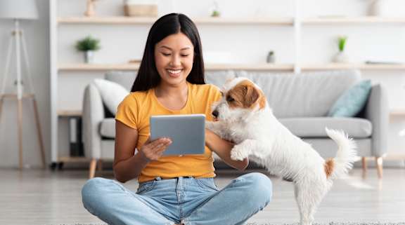 A woman is sitting on the floor with a tablet, and a small dog is standing on its hind legs beside her.