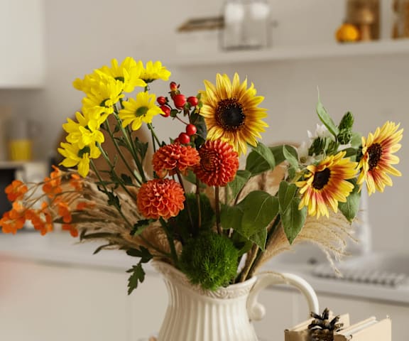 A white vase with sunflowers and other flowers on a kitchen counter.