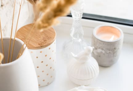 A white ceramic candle holder with a wooden lid is on a table next to a white ceramic candle holder with a marbled pattern and a white ceramic bird figurine.