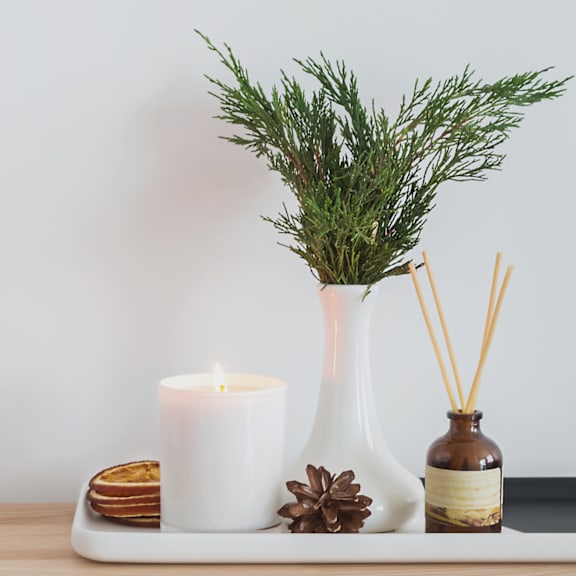 A white candle, a pine branch, and a brown bottle of reed diffuser on a wooden tray.