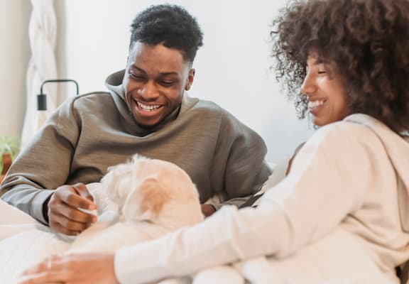A man and woman are sitting on a bed with a dog between them.