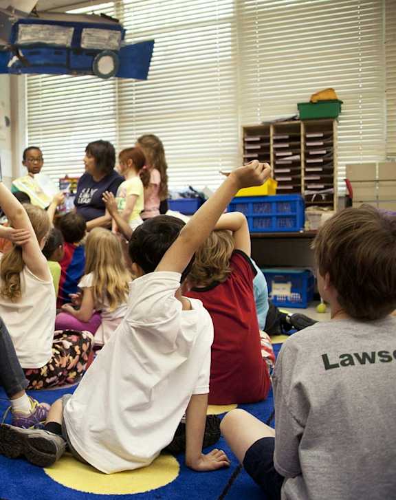 A group of children are sitting on the floor in a classroom with their hands raised.