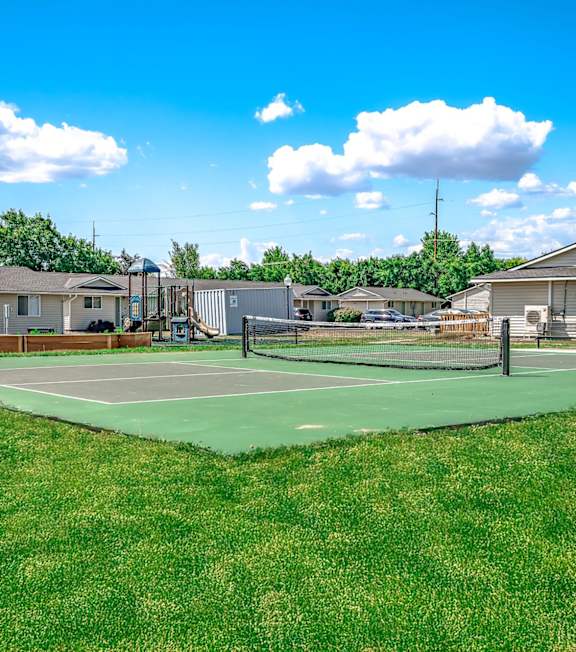 A tennis court is surrounded by grass and houses.