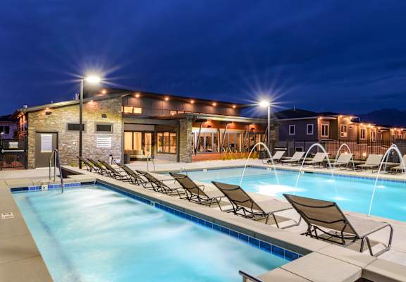 A swimming pool with lounge chairs and lights on at night.