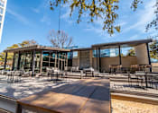 Thumbnail 5 of 24 - A modern building with a glass facade and a wooden deck with chairs and tables.