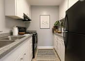 Thumbnail 6 of 8 - A black refrigerator sits in a kitchen with white cabinets and a grey counter.