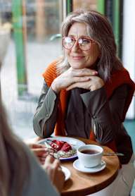 A woman with glasses and a scarf is sitting at a table with a cup of coffee and a plate of food in front of her.