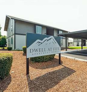A sign for Dwell at Life Apartment Homes stands in front of a building.