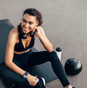A woman in workout gear sits on a mat with a water bottle, a soccer ball, and headphones.