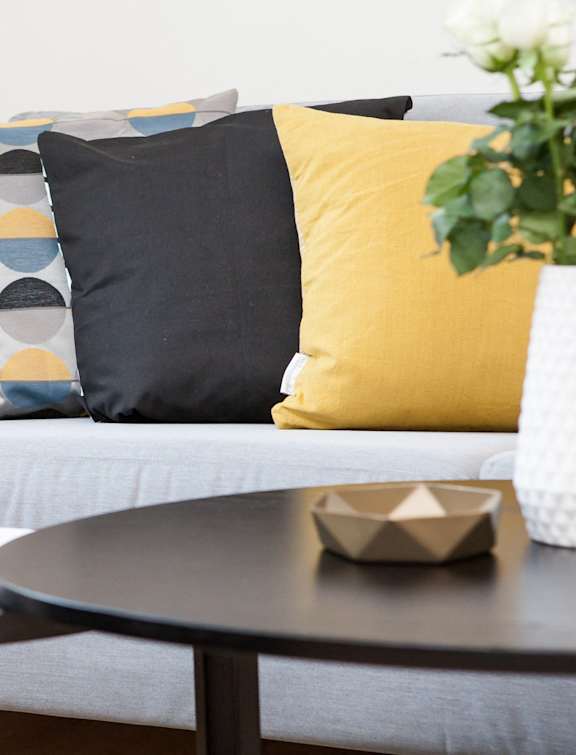 A grey couch with a yellow pillow and a vase with white flowers on a table.
