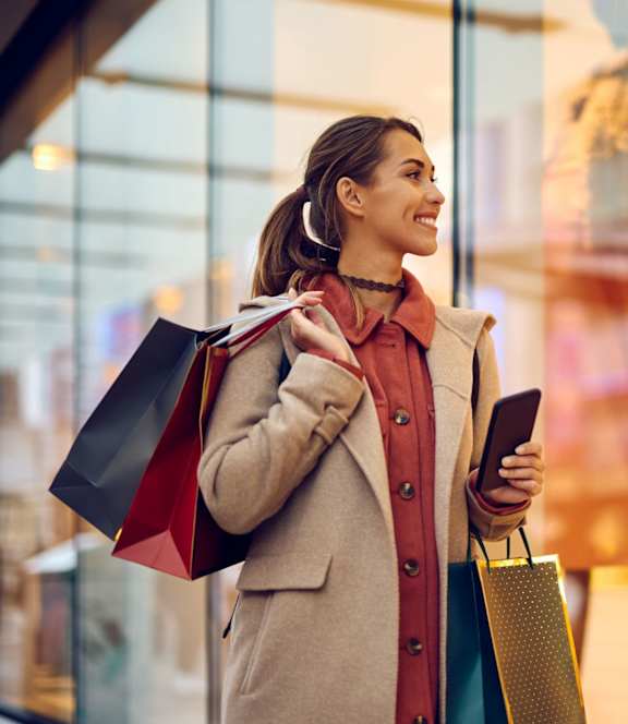 A woman is standing outside a store holding shopping bags and a phone.