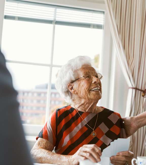 an elderly woman sits at a table in front of a window