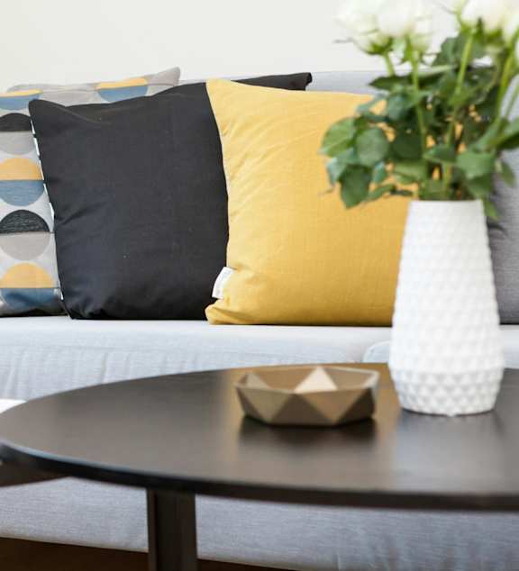 A yellow mug sits on a coffee table in front of a grey couch with a yellow pillow and a white blanket.