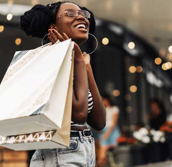 Kerrville, TX, Apartments Near River Hills Mall - Winwood Club - Neighborhood Shopping Scene With Smiling Resident Holding Store Bags, Casual Style, and Soft String Lights Along A Pathway.