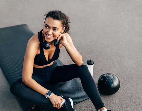 A woman in workout gear sits on a mat with a water bottle, a soccer ball, and headphones.